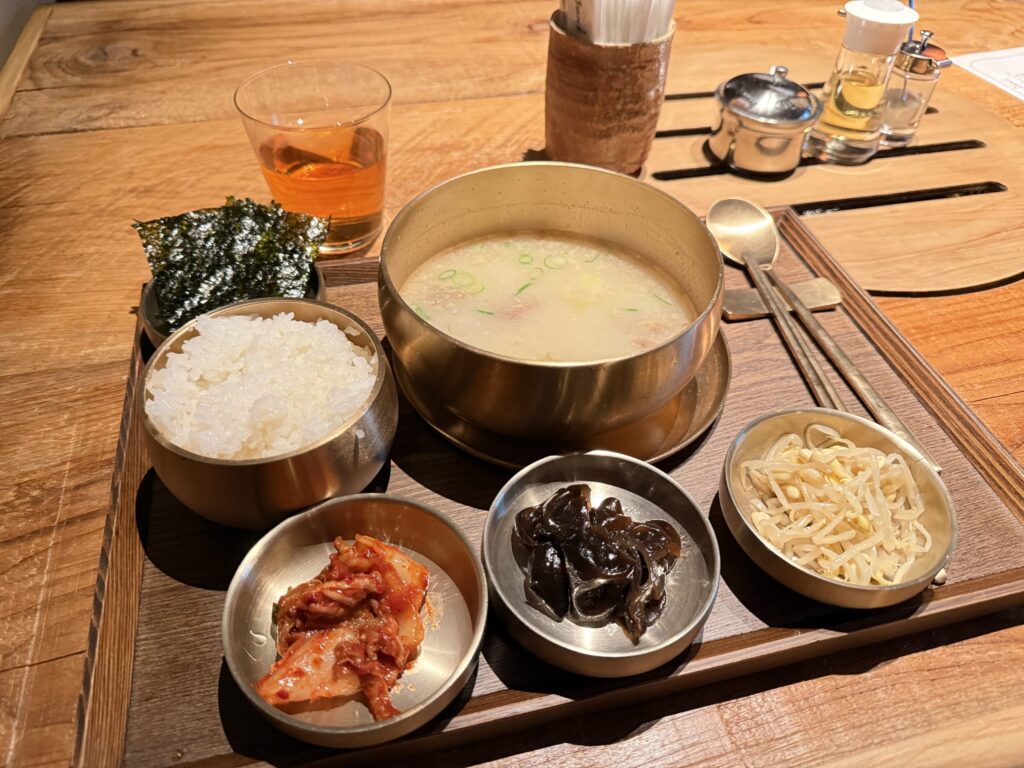 木のトレーに並べられた和牛骨湯（コムタン）の定食セット。真鍮の器に入った白濁スープ、ご飯、キムチ、海苔、ナムルなどの小鉢。