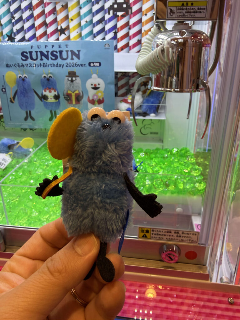 A hand holding a blue Puppet Sunsun plush mascot with a yellow balloon in front of a claw machine filled with green plastic gems