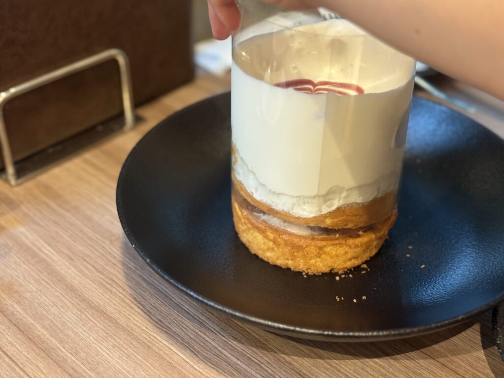 Close-up of a hand lifting a clear plastic film from a stack of pancakes, releasing a cascade of white cream and berry sauce on a black plate
