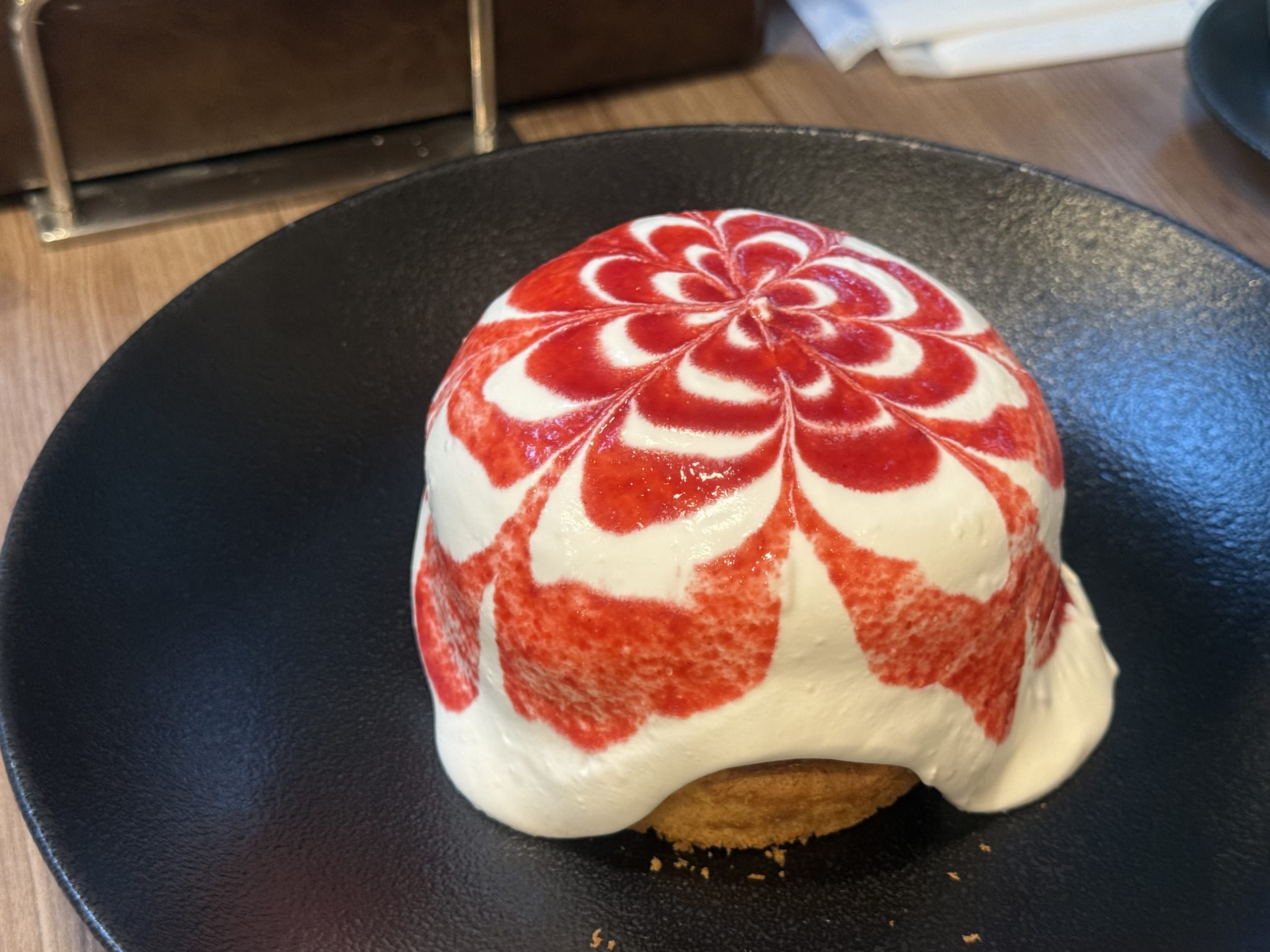 A pancake completely covered in white cream with a red strawberry sauce flower pattern, served on a black plate