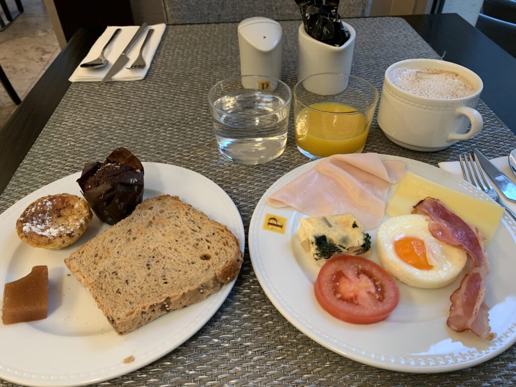 A lavish breakfast table spread featuring Pastel de Nata, toast, muffins, ham, fried eggs, bacon, cappuccino, and orange juice