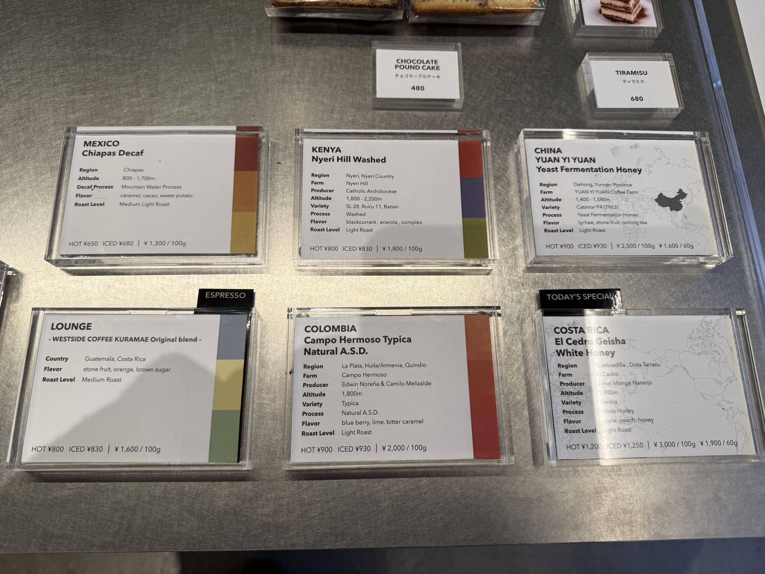 Coffee bean menu cards displayed on a metal counter, featuring various single-origin options like Mexico, Kenya, China, Colombia, and Costa Rica with prices and flavor notes