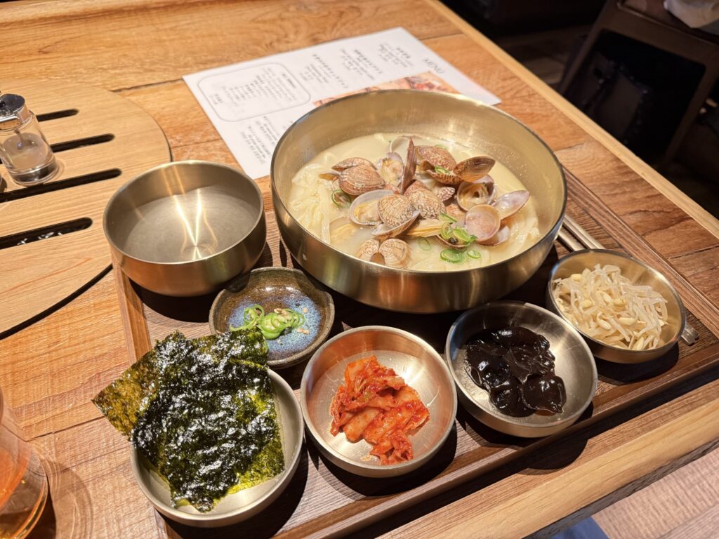 A set meal featuring Kalguksu noodle soup topped with abundant clams in a brass bowl, served with kimchi, seaweed, and other side dishes