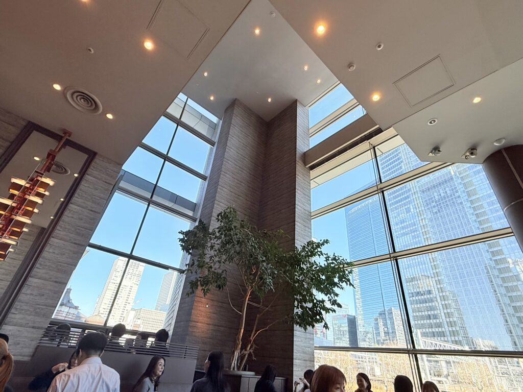 Spacious interior with high ceilings and large windows overlooking city skyscrapers and blue sky, with people seated at tables
