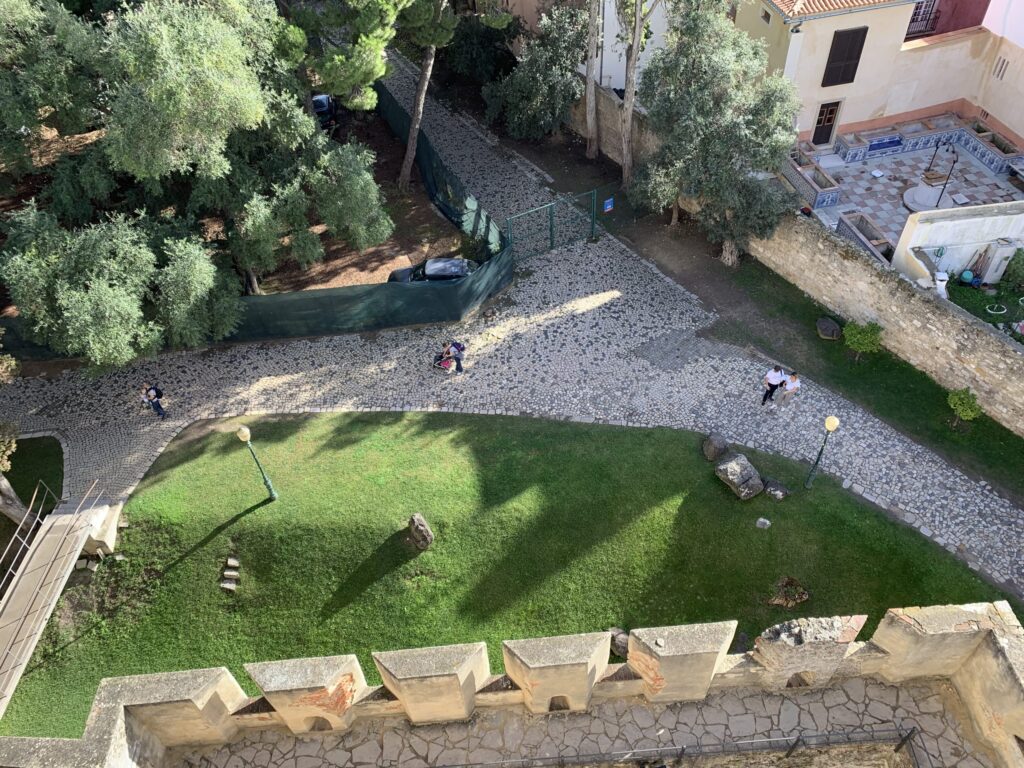 High-angle view from a castle wall looking down at a courtyard with geometric cobblestone paths and a green lawn