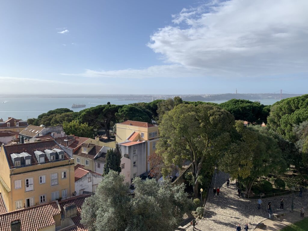 View from the castle walls overlooking lush green trees, traditional rooftops, the Tagus River, and the 25 de Abril Bridge in the distance