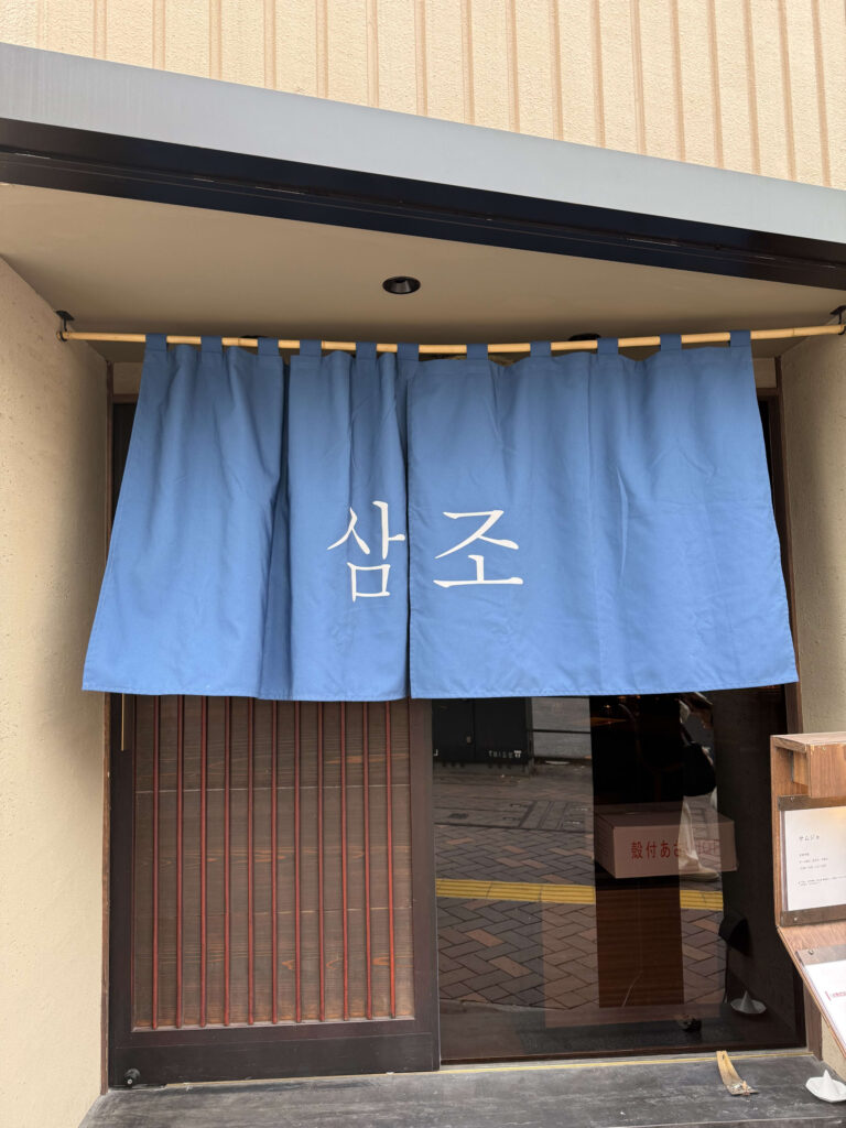 A bright blue noren curtain hanging at a shop entrance with white Korean text reading Samjo