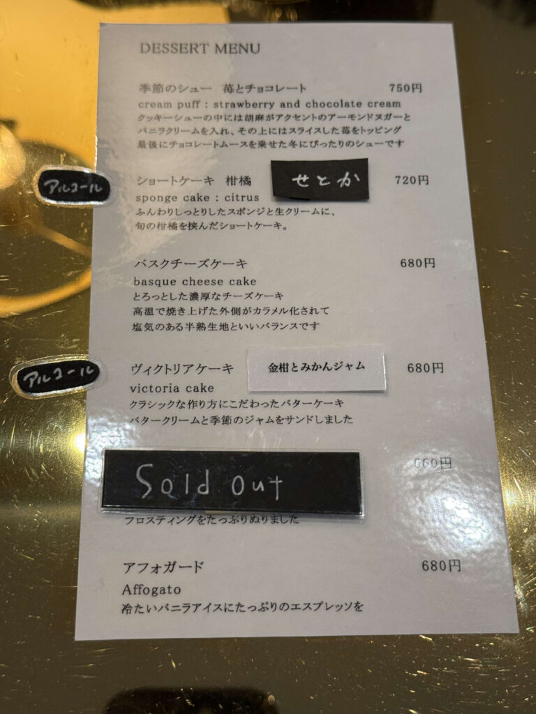 Dessert menu sheet on a gold table listing items like Basque Cheese Cake, Victoria Cake, and seasonal shortcakes with prices.