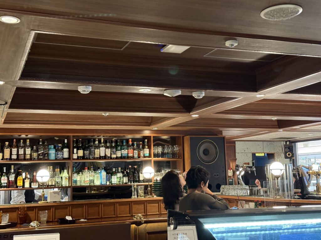木製の棚にずらりと並ぶ洋酒ボトルと大きなスピーカーが特徴的な、レトロモダンなバーの店内の様子