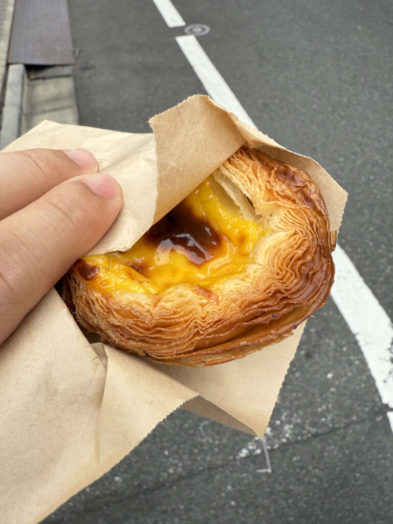 Close-up of a hand holding a freshly baked egg tart wrapped in brown paper against a street background