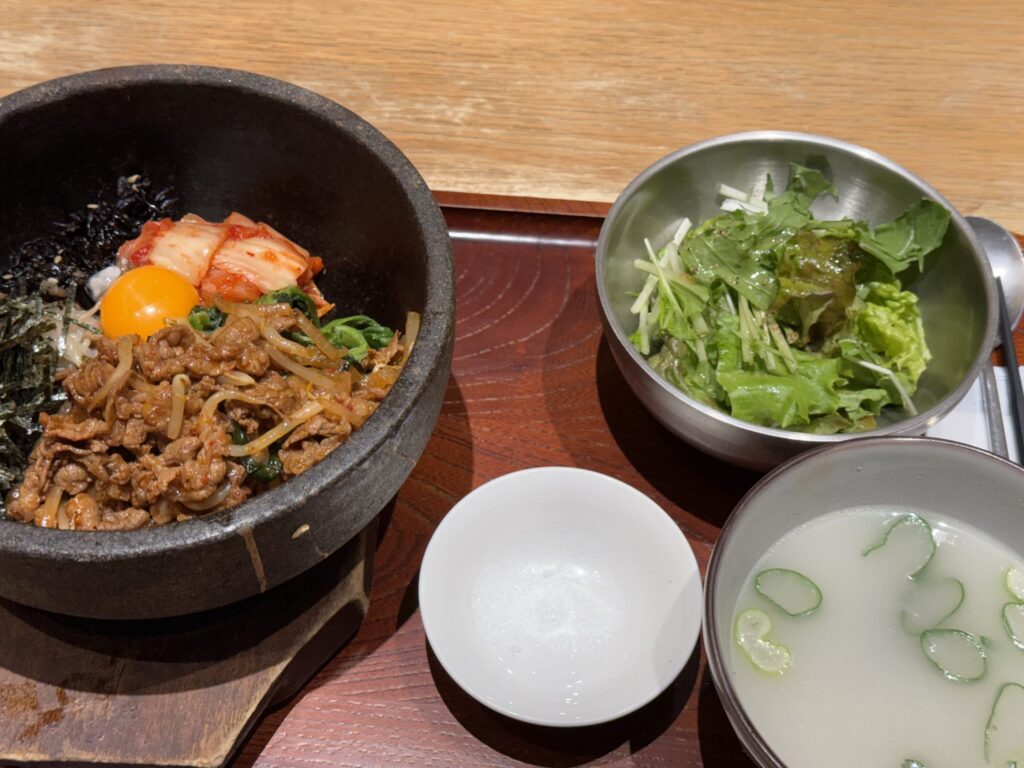 Jeonju Dolsot Bibimbap set meal featuring a hot stone bowl rice dish, green salad, and milky white Seolleongtang soup on a wooden tray.