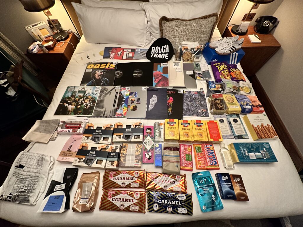 A massive haul of UK souvenirs spread out on a hotel bed, including vinyl records, snacks, cosmetics, and sneakers, all purchased thanks to a backup VISA card.