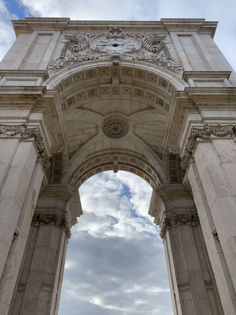 Low-angle view looking up at the Arco da Rua Augusta in Lisbon, featuring intricate stone carvings, a clock, and the coffered archway against a cloudy sky