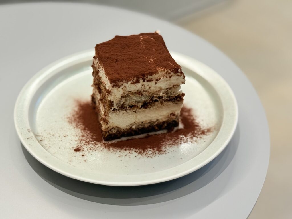 A square slice of tiramisu covered in cocoa powder served on a round white plate