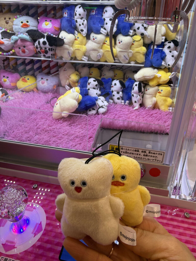 A hand holding two small plush toys, a cream bear and a yellow duck by mojojojo, in front of a claw machine
