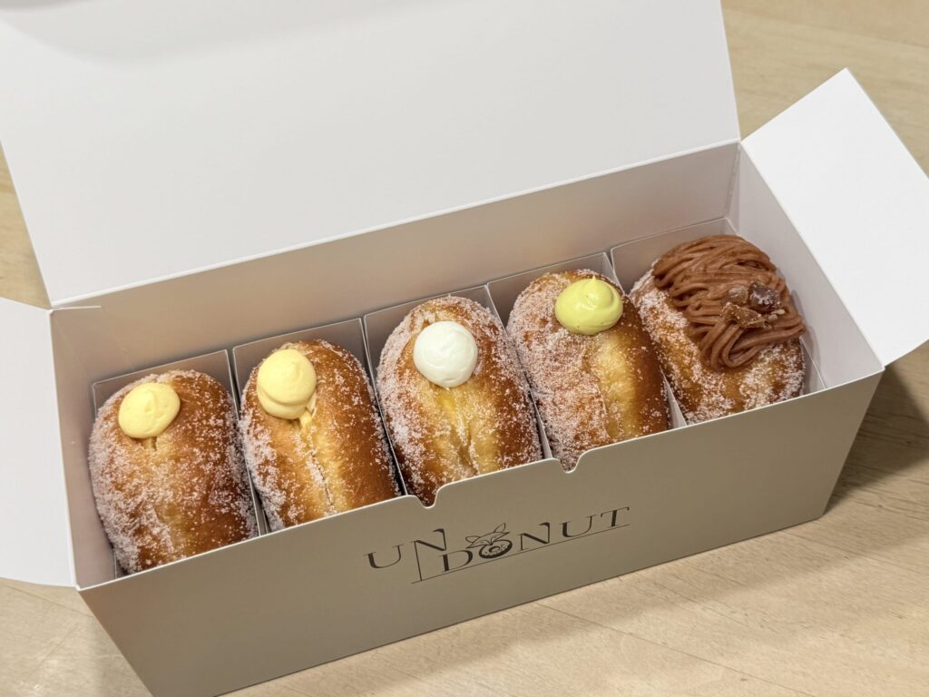 Five sugar-coated cream donuts arranged in a white UN DONUT box, featuring custard, whipped cream, pistachio, and Mont Blanc flavors.