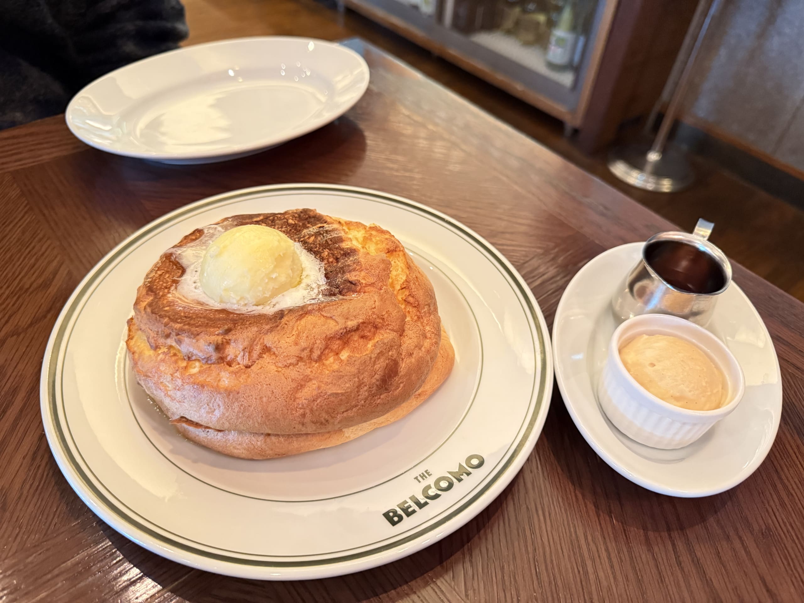 A thick, golden-brown pancake on a plate with THE BELCOMO logo, topped with a scoop of butter and served with side toppings