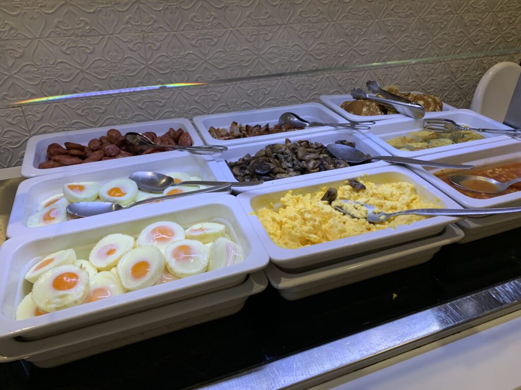 ホテルの朝食ビュッフェにあるホットミールコーナー。目玉焼き、スクランブルエッグ、ソーセージ、ベーコン、マッシュルームなどが白いトレイに並んでいる様子