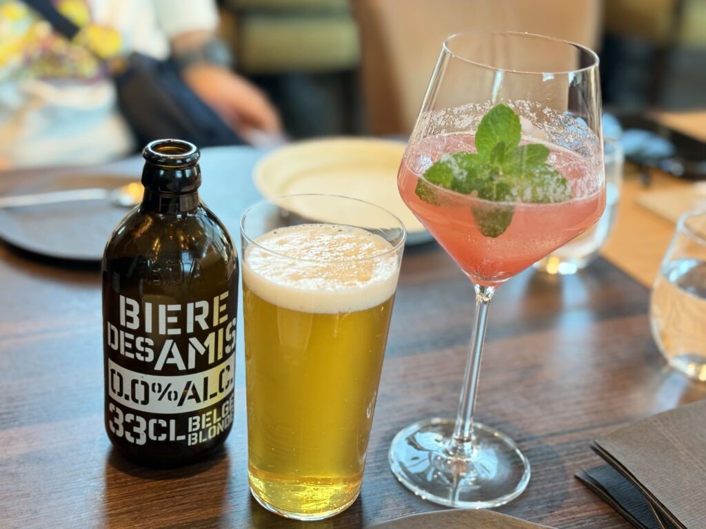 テーブルに置かれたビア・デザミ・ブロンドのノンアルコールビール瓶とグラス、およびミントが添えられたピンク色のグァバ・ライムのモクテル
