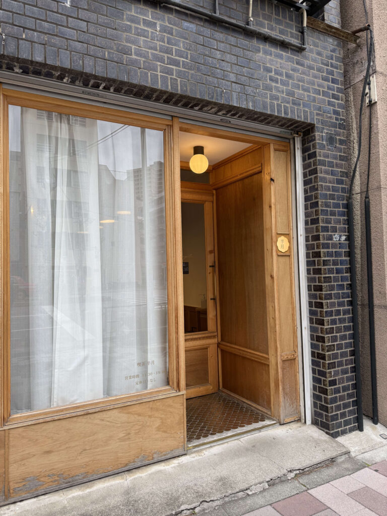 Exterior of 'Kissa Hangetsu' featuring a dark brick facade, large wooden window frames with white curtains, and a warm pendant light