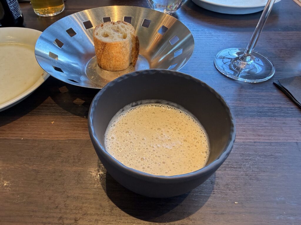 Foamy Topinambur (Jerusalem artichoke) potage in a dark bowl with a slice of baguette in a metal basket behind it