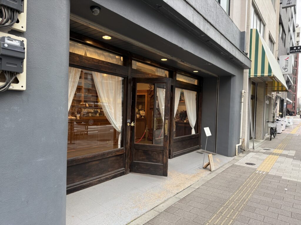 Exterior of a shop featuring heavy wooden doors, white curtains in the windows, and grey walls