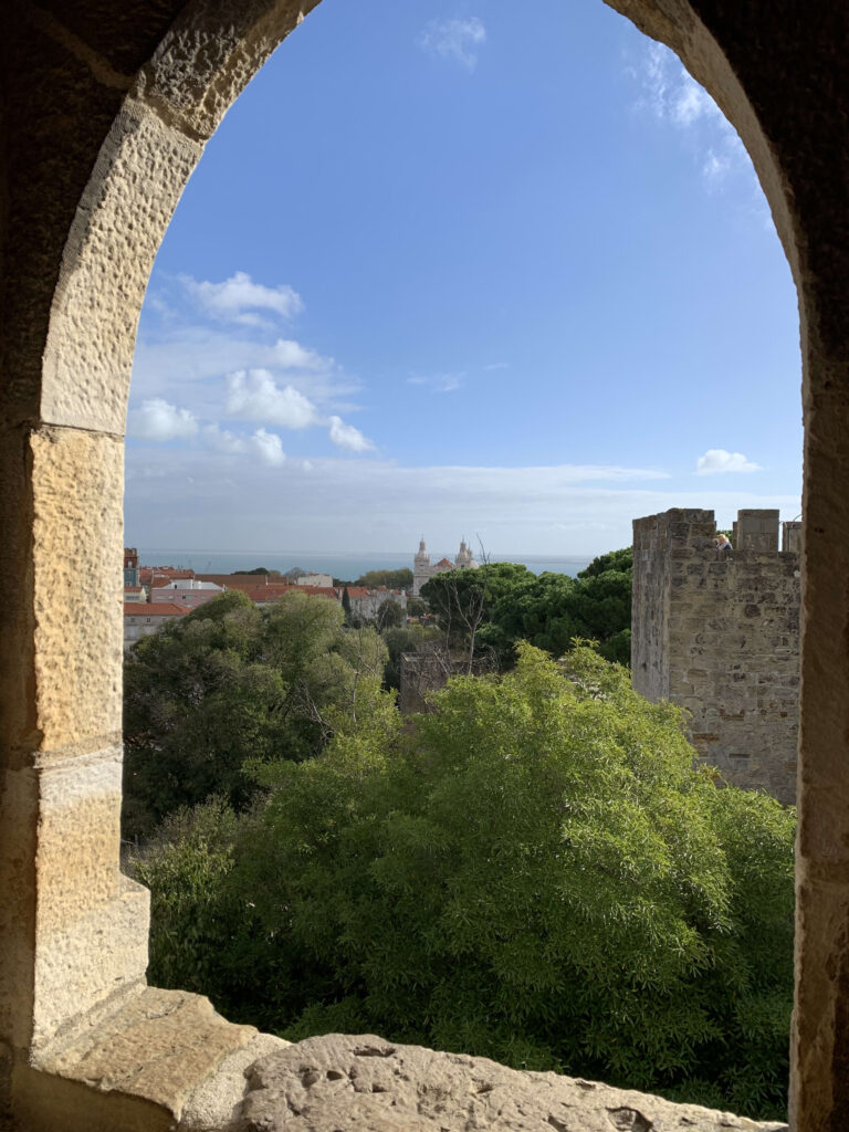 A scenic view of lush trees, a distant white church, and blue sky framed by a rustic stone archway of a castle