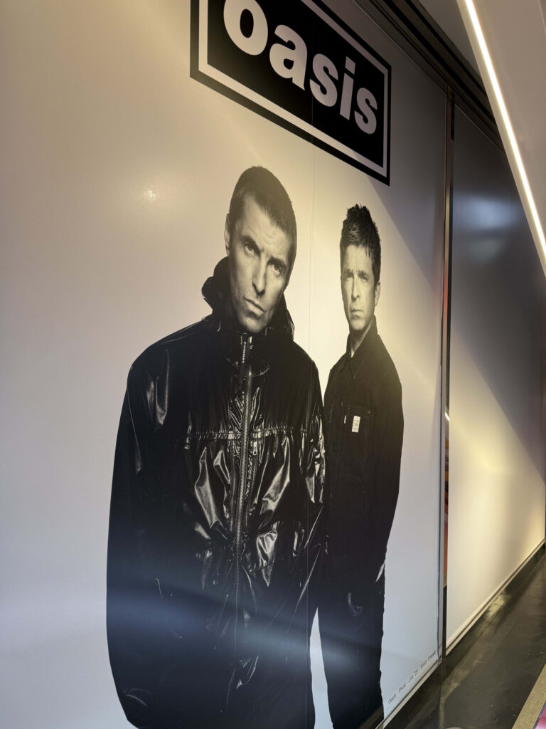 Large wall advertisement for Oasis featuring a black and white portrait of Liam and Noel Gallagher under the band's logo