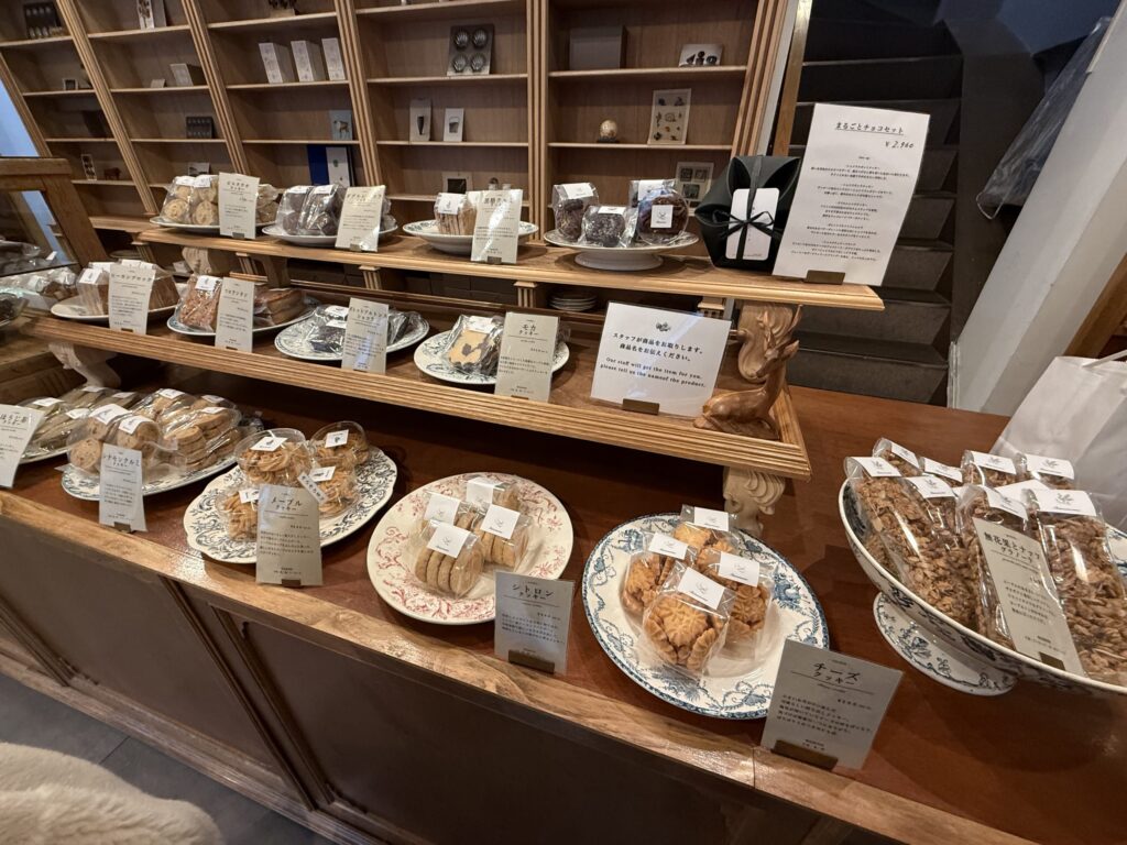 木製の陳列棚とカウンターに並べられた豊富な種類の焼き菓子。シナモンくるみクッキー、メープルクッキー、シトロンクッキーなどが皿に盛られ、ギフト用のチョコセットも見える。