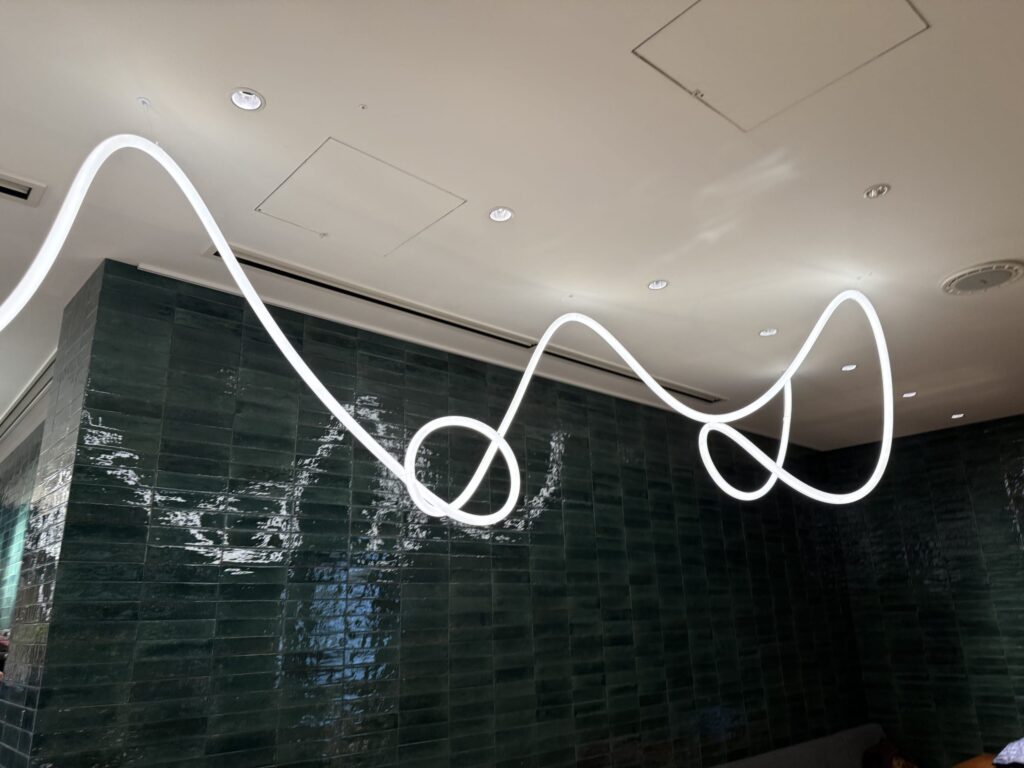A modern white tube light fixture hanging from the ceiling in wavy loops against a dark green glossy tiled wall