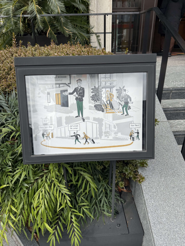 A black-framed illustrated sign standing in a planter outside The Aoyama Grand Hotel. The cartoon map guides visitors to a 'more accessible entrance nearby' to avoid the stairs, showing a concierge character pointing the way.