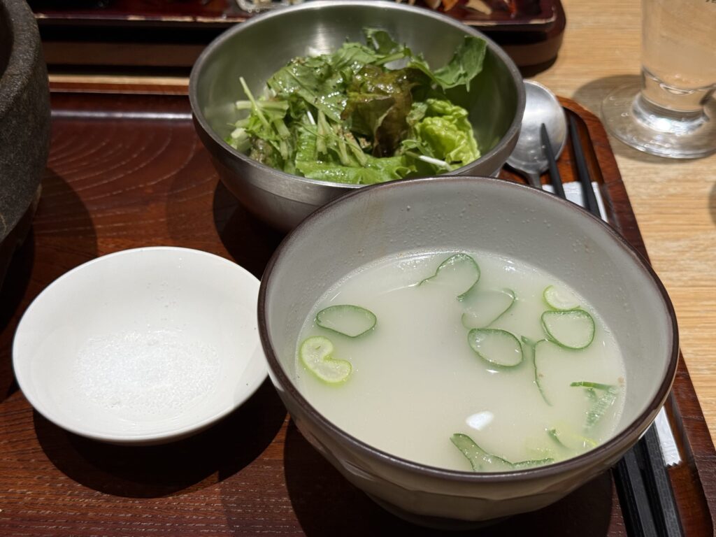 Milky white Seolleongtang soup with green onions, a small dish of salt, and a green salad in a metal bowl on a wooden tray