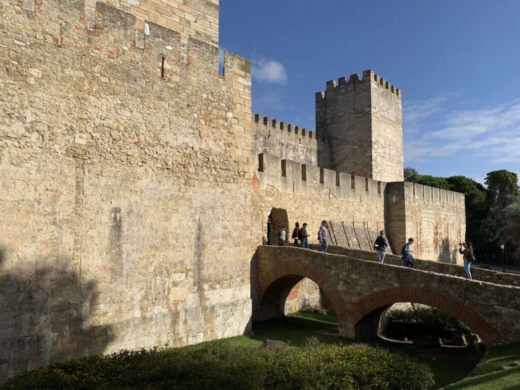 青空の下、堅牢な中世の要塞の城壁と、内郭へ続く石橋を渡る観光客