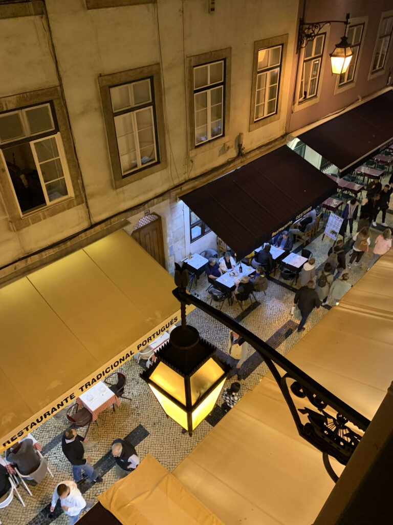 High-angle view of a cobblestone street at night featuring a Portuguese restaurant with a yellow awning and outdoor dining tables
