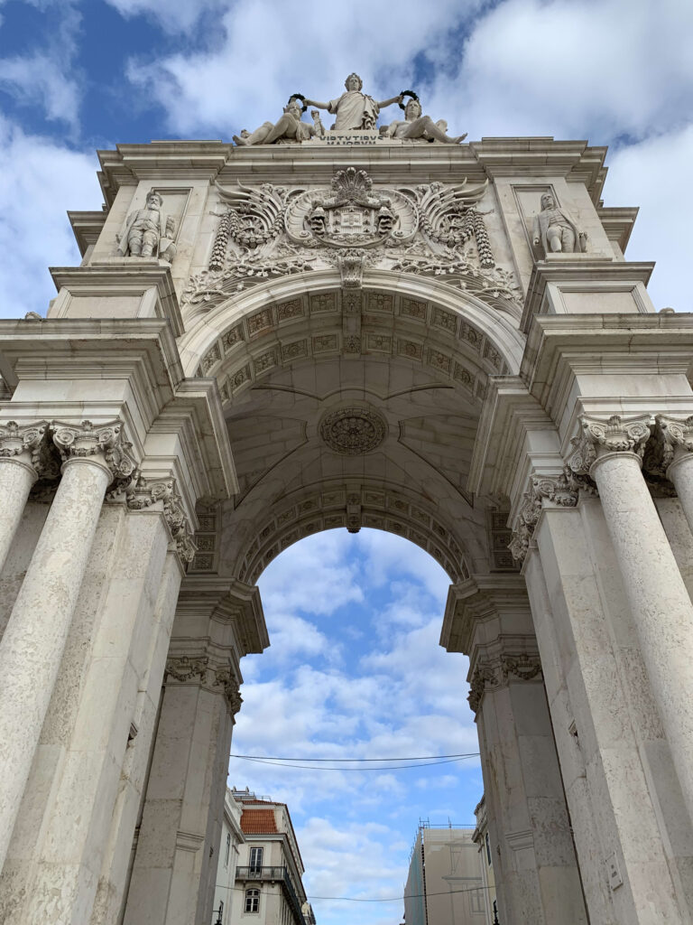 Low-angle view of the Rua Augusta Arch in Lisbon featuring the statues of Glory, Genius, and Valor against a blue sky