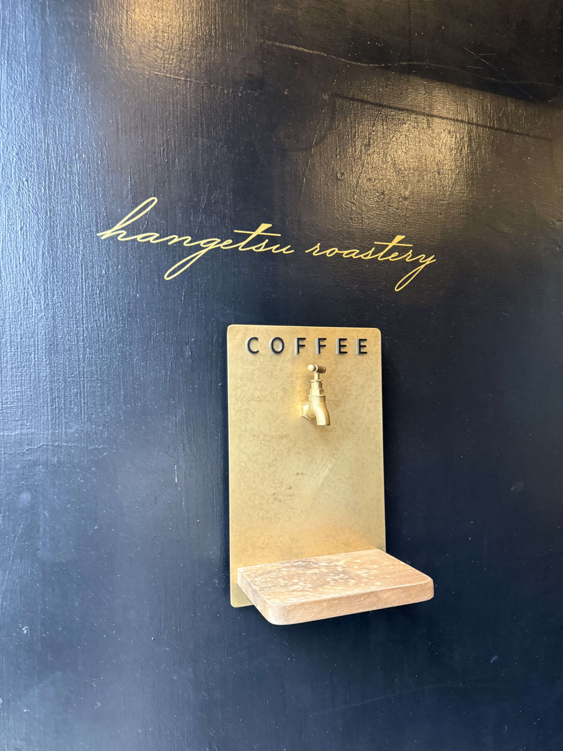濃紺の壁面に描かれた「hangetsu roastery」の金色の筆記体ロゴと、「COFFEE」の文字が入った真鍮製の蛇口型オブジェ