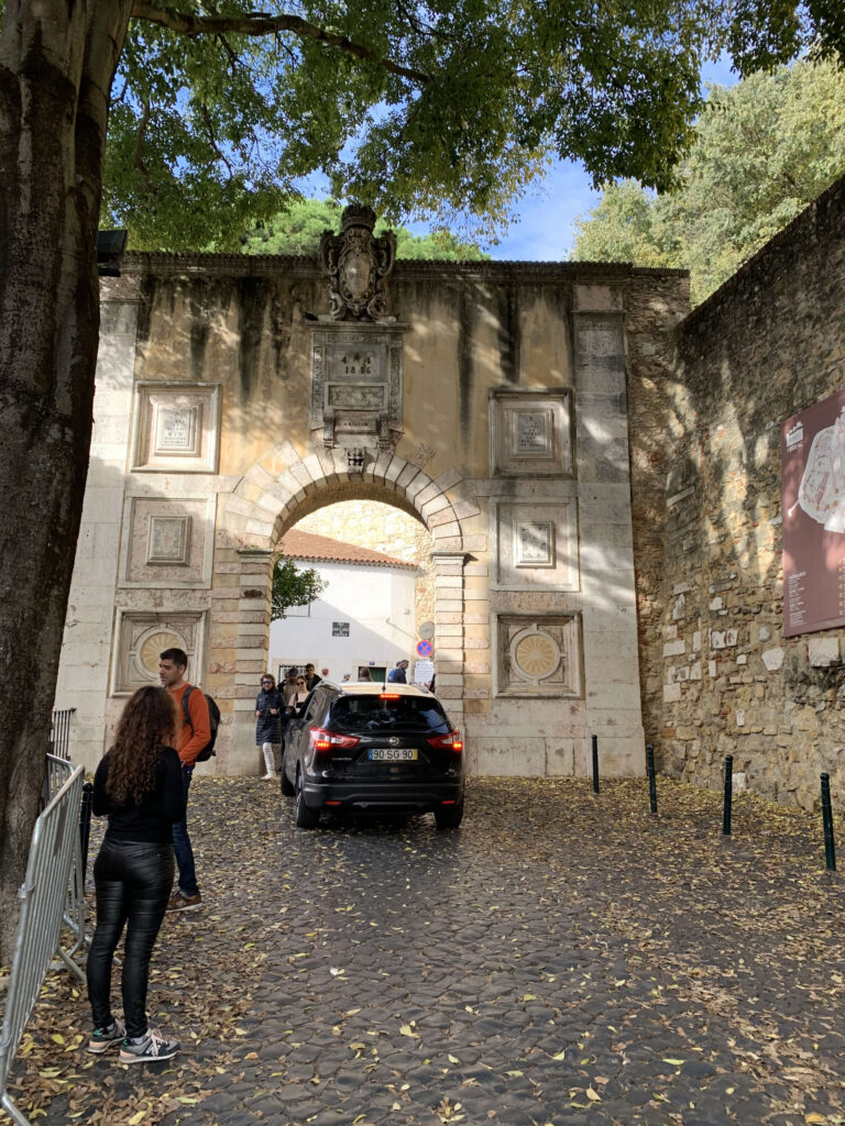 A black car driving through the Arco do Castelo, the 19th-century stone archway entrance to St. George's Castle, with visitors standing on the cobblestone path.