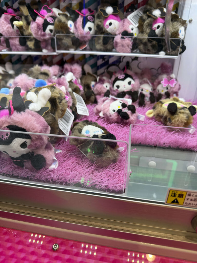Sanrio plush toys including Kuromi and Cinnamoroll wearing leopard print hoods displayed inside a claw machine on pink fur