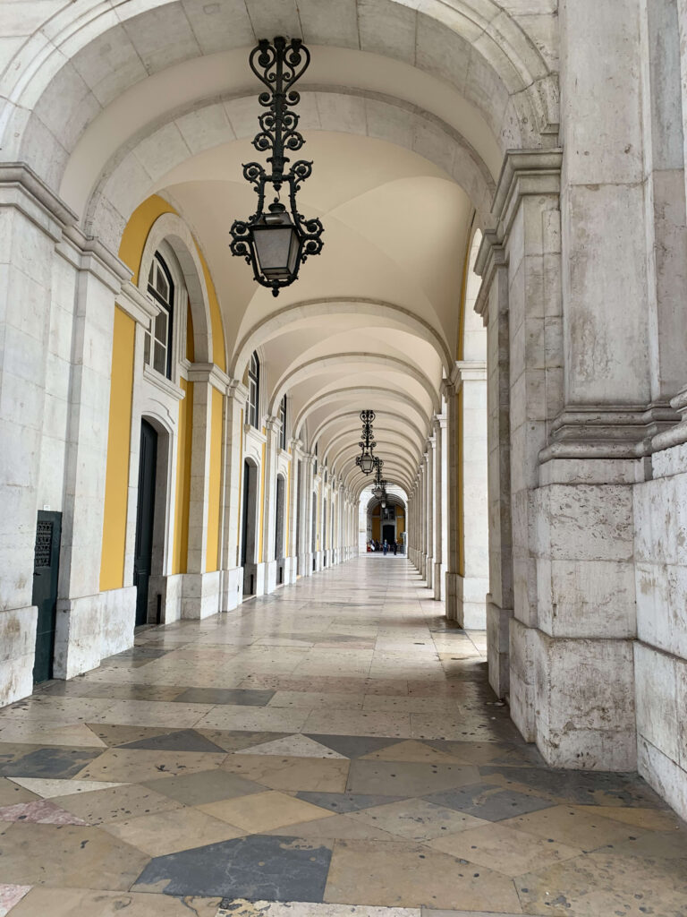 A long arcade corridor in Lisbon featuring mustard yellow walls, repeating white stone arches, and hanging black metal lanterns