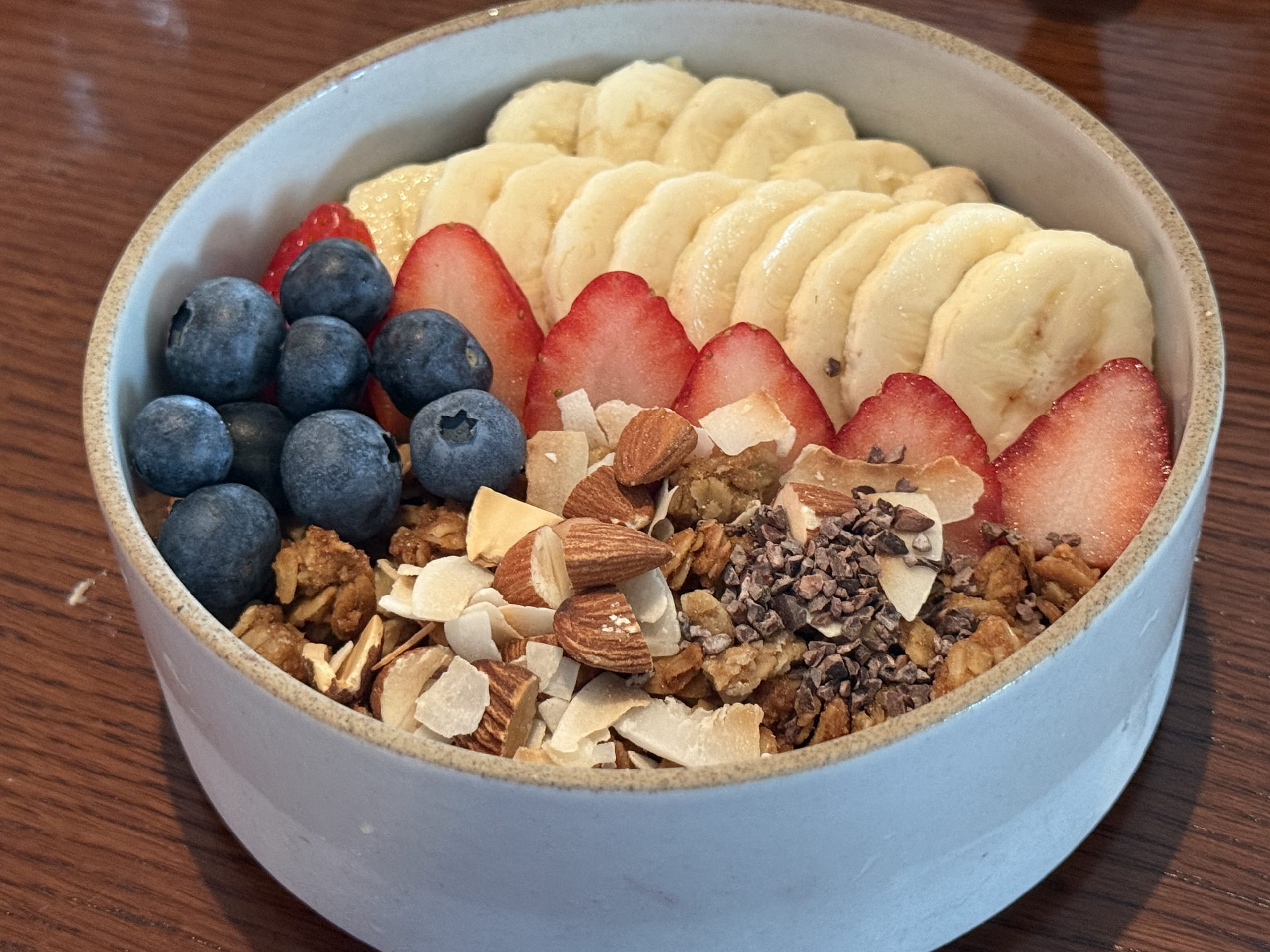 A ceramic bowl filled with a smoothie base topped with blueberries, sliced strawberries, bananas, almonds, coconut flakes, cacao nibs, and granola on a wooden table.