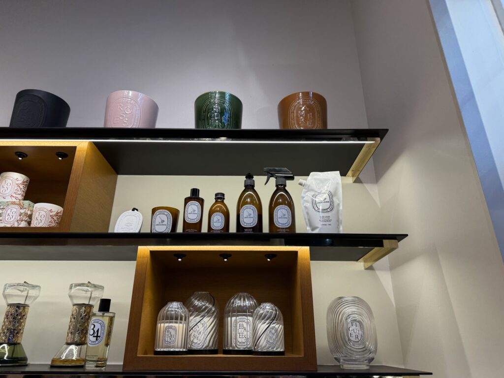 Display shelves featuring Diptyque products: large colored candles on top, La Droguerie home care collection in the middle, and hourglass diffusers with glass-domed candles on the bottom.