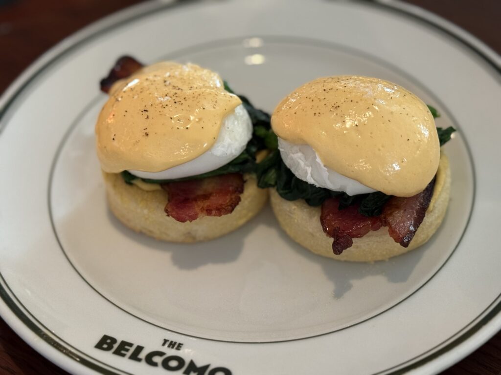 Two eggs benedict served on a white plate with 'THE BELCOMO' logo, featuring English muffins topped with bacon, spinach, poached eggs, glossy hollandaise sauce, and black pepper.