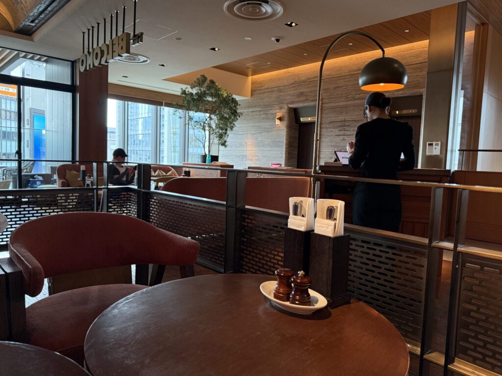 Interior of The Belcomo restaurant at Aoyama Grand Hotel, featuring a wooden table with condiments in the foreground, terracotta velvet chairs, a staff member at a counter, and large windows overlooking the city