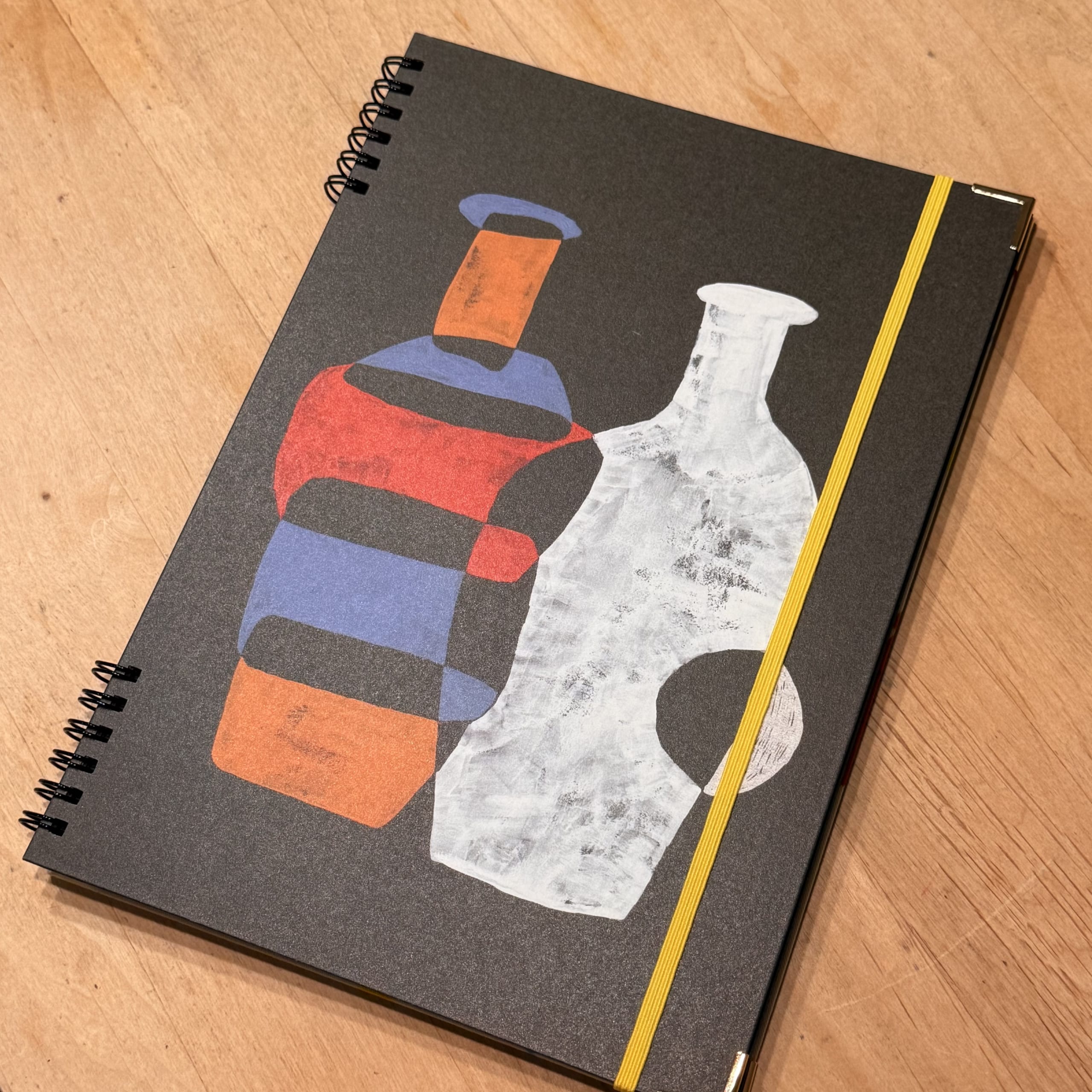 A black spiral notebook with painted bottles on a desk.