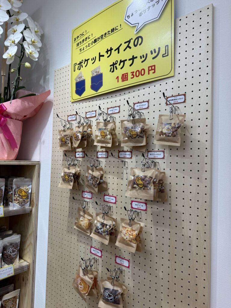 A shop display featuring a yellow sign that reads 'Pocket-sized Pokenuts' on a pegboard. Small individual bags of nuts and dried fruits are hanging by clips arranged in rows, with a white orchid and wooden shelf visible on the left.