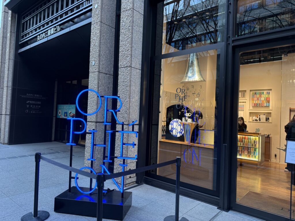 A bright blue neon sign sculpture spelling ORPHÉON standing in front of the Marunouchi Nijubashi Building