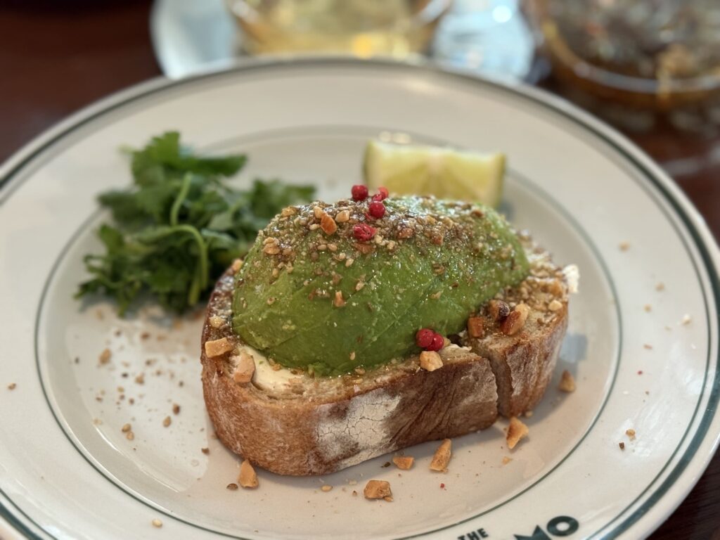 Avocado toast on a white plate with a green rim, topped with a whole avocado half, crushed nuts, spices, and pink peppercorns, served with fresh cilantro and a lime wedge.