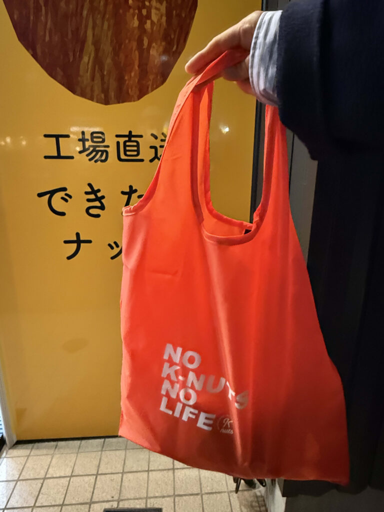 「NO K-NUTS NO LIFE」とプリントされたオレンジ色のエコバッグを持つ手と、背景の「工場直送」と書かれた黄色い看板