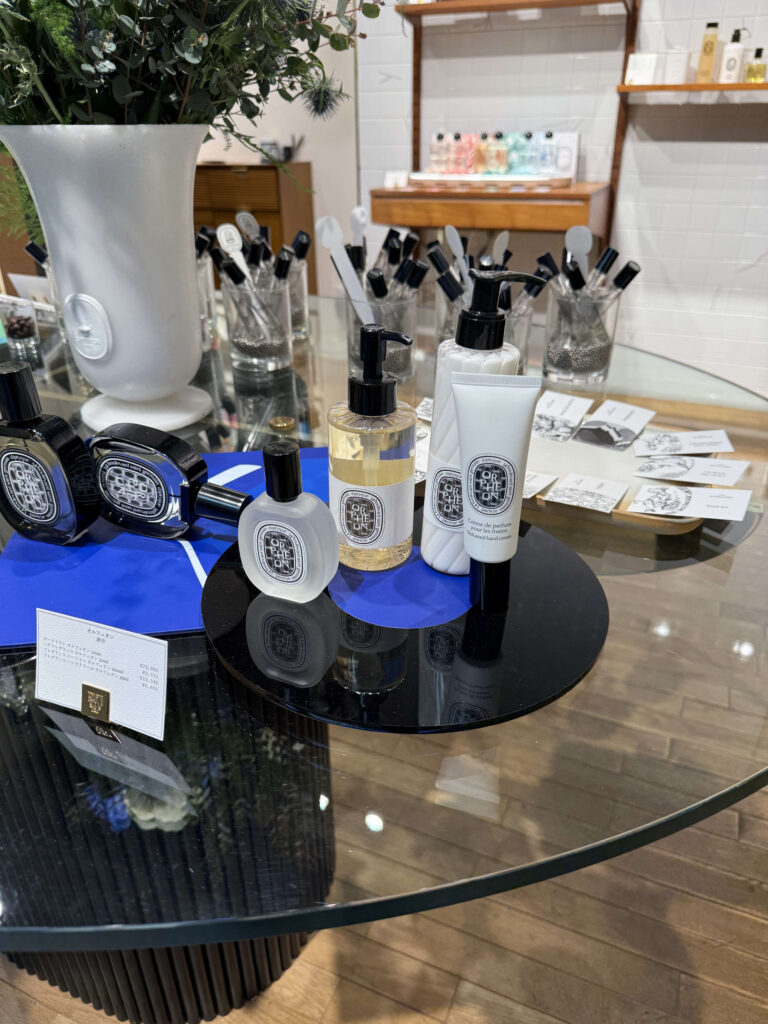 Diptyque Orphéon collection display featuring eau de parfum, hand cream, and hair mist with a price list on a glass table
