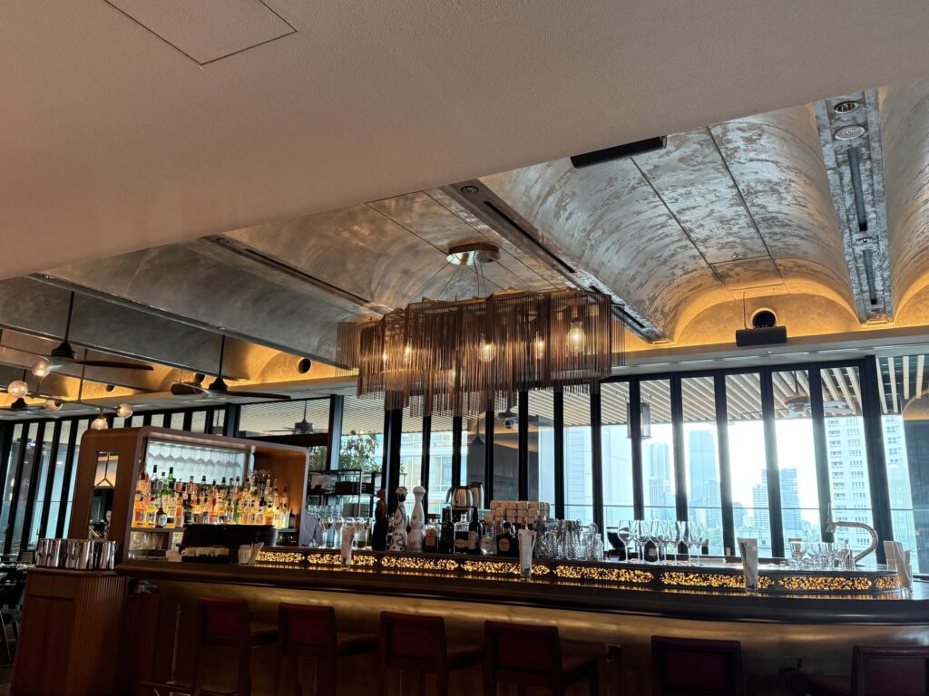 Interior of a modern luxury bar featuring an arched concrete ceiling, metallic fringe chandelier, and floor-to-ceiling windows overlooking a cityscape